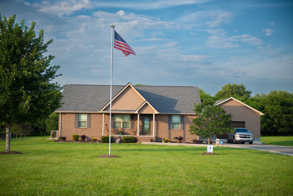 American flag in front of a brick house with a spacious lawn and a car parked in the driveway.