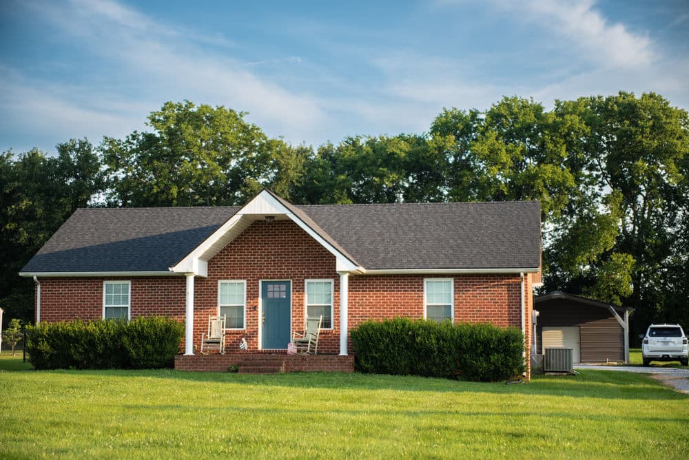 Brick house with blue door, green lawn, and trees in the background on a sunny day.