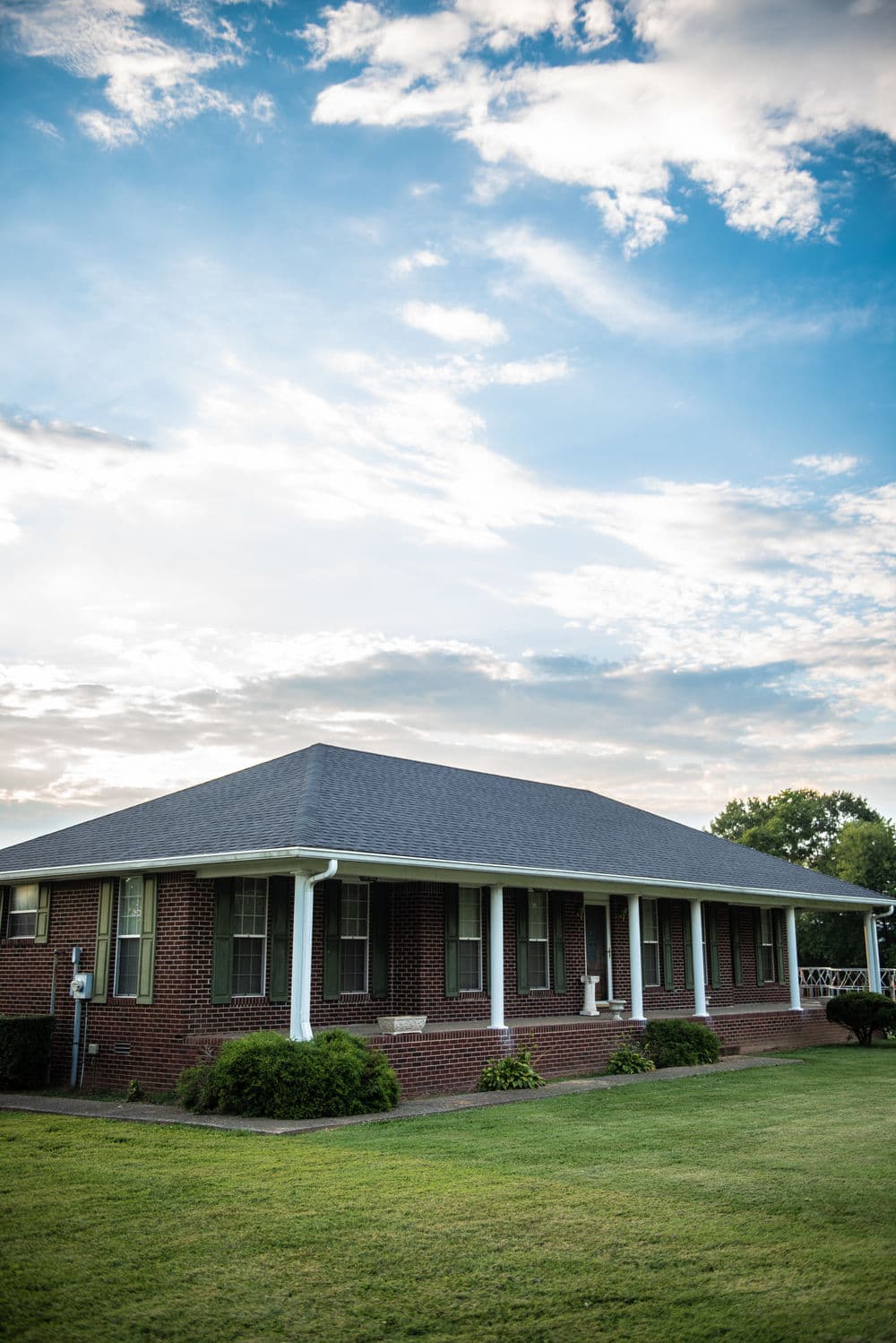Brick house with a porch surrounded by green lawn under a blue sky with scattered clouds.