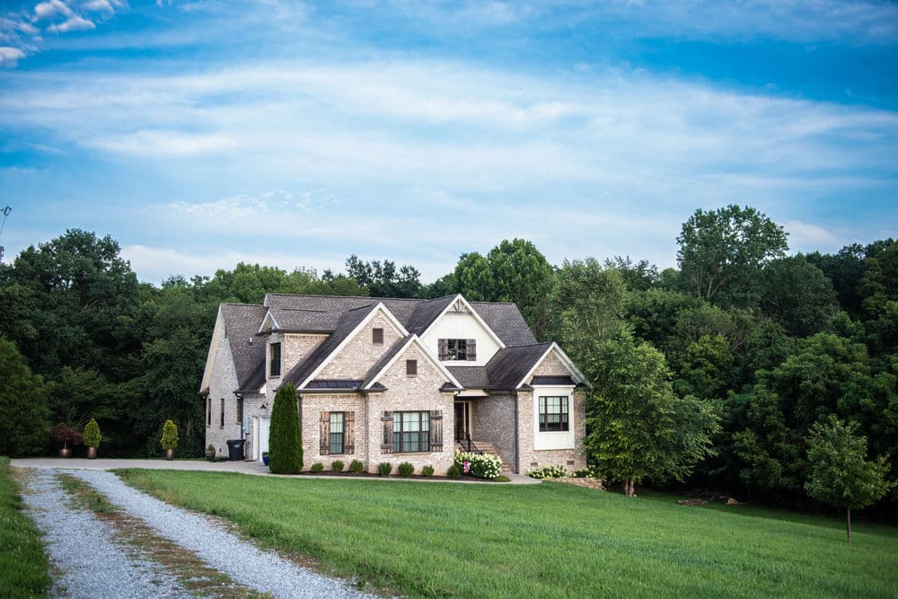 Modern stone house surrounded by trees and a gravel driveway under a blue sky.